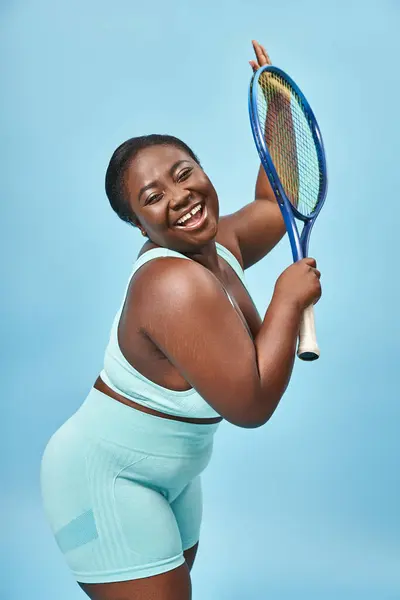 又大又黑的皮肤女人拿着蓝色背景的网球拍 身体正面和运动 — 图库照片