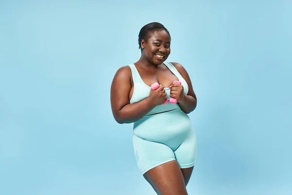 happy plus size woman in blue active wear exercising with dumbbells on matching background