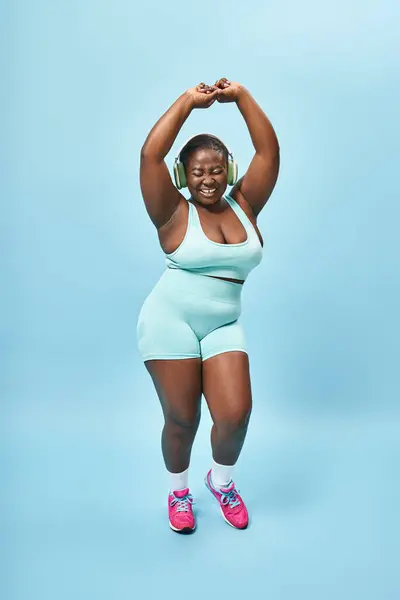 happy plus size african american woman dancing in active wear with headphones on, blue background