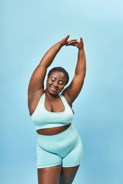 excited plus size african american woman in active wear stretching joyfully on blue background