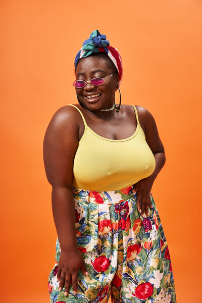 cheerful african american plus size woman with floral skirt and sunglasses posing on orange backdrop