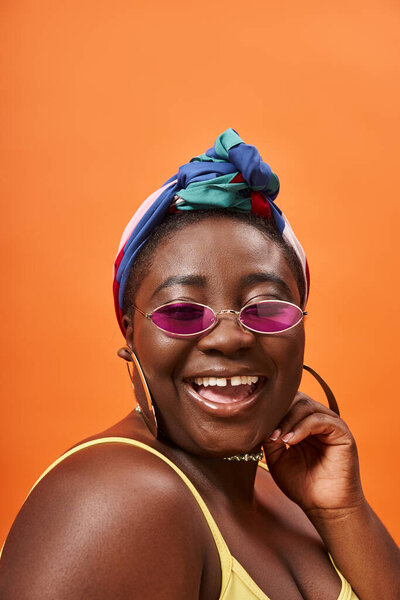 cheerful and plus size african american woman in headscarf and trendy sunglasses on orange