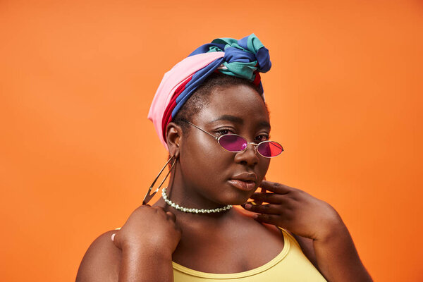Fashionable plus size and young african american woman in headscarf and trendy sunglasses on orange