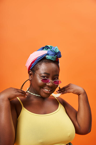 Fashionable plus size and happy african american woman in headscarf and trendy sunglasses on orange