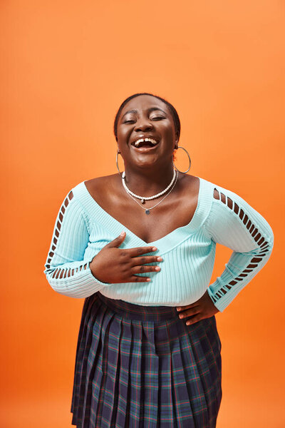 cheerful plus size african american woman in blue long sleeve smiling and posing with hand on hip
