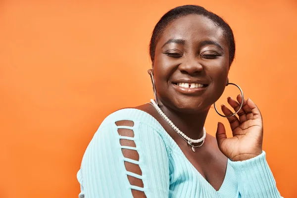 stock image portrait of pleased plus size african american woman in blue long sleeve smiling on orange