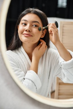 young asian woman with brunette hair using jade roller for a facial massage in bathroom, anti-aging clipart