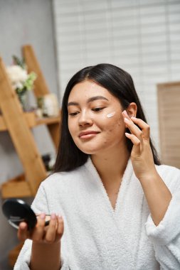 Esmer saçlı ve aynaya bakan Asyalı genç bir kadının yüzünde sivilce kremi.