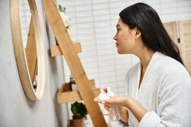 Modern banyoda esmer saçlı, yüz temizleyici köpük tutan Asyalı genç bir kadının yan görüntüsü.