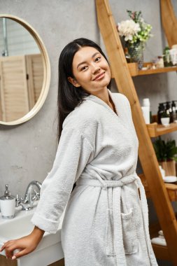 Güzel ve genç Asyalı kadın banyo bornozuyla modern banyoda kameraya bakıyor.