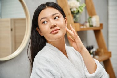 Banyo bornozu giymiş genç Asyalı kadının portresi sivilceye yatkın tenine dokunuyor ve banyoya bakıyor.