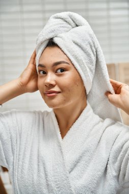Kafasında beyaz havlu olan güzel Asyalı kadın banyoda kameraya bakıyor, deri sorunları var.