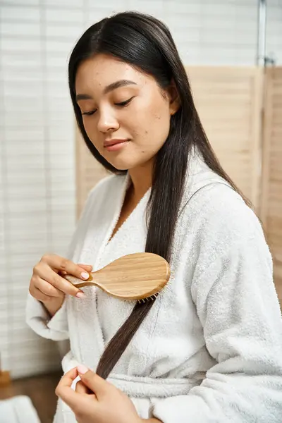 young asian woman in robe combing her brunette healthy hair with wooden hairbrush in bathroom