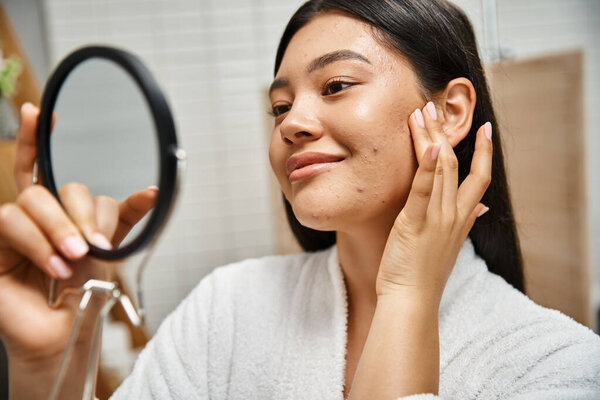 young and brunette asian woman with pimples examining her face in mirror, skin issues of real people