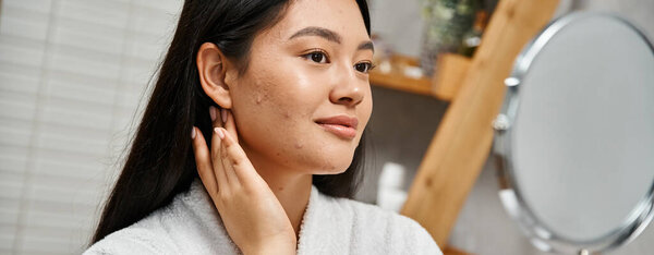 portrait of brunette and young asian woman with acne looking at mirror in modern bathroom, banner