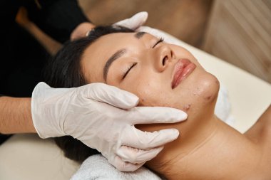 facial treatment for acne-prone skin, masseuse in latex gloves and asian woman with closed eyes clipart