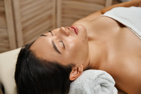 young and brunette asian woman with closed eyes lying in wellness center, spa and facial treatment