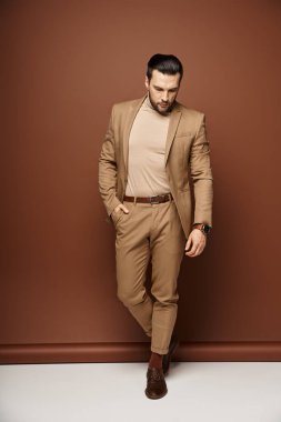 handsome man in elegant attire looking down while posing with hand in pocket on beige background clipart