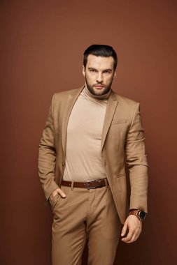 handsome man in elegant attire looking at camera while posing with hand in pocket on beige backdrop clipart