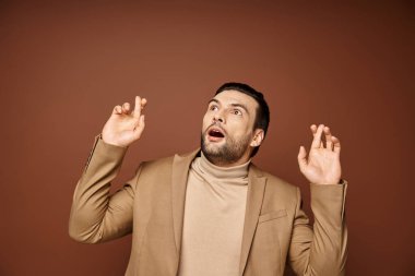 astonished man in elegant attire with his fingers crossed looking up on beige background clipart