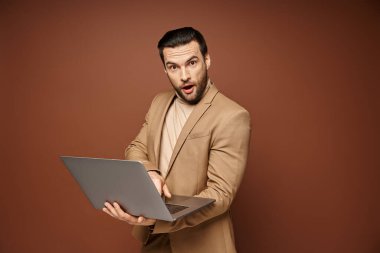 Şok olmuş ve yakışıklı profesyonel bilgisayarını kullanarak bej arka plan üzerinde çalışıyor.