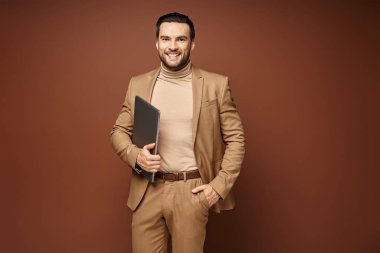 Şık giyinmiş mutlu iş adamı bej arka planda dizüstü bilgisayarıyla duruyor.