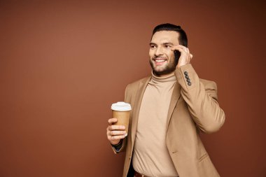 Havalı giyinmiş, elinde kahve tutan mutlu bir adam bej arka planda akıllı telefondan arıyor.