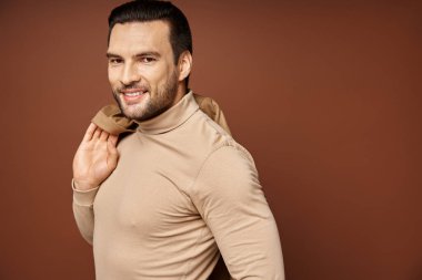 Balıkçı yakalı mutlu bir adam. Omzunda modaya uygun bir ceket tutuyor ve bej renkli kameraya bakıyor.