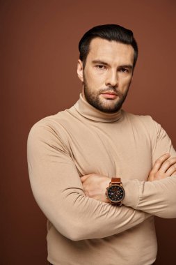 Balıkçı yakalı kararlı ve yakışıklı bir adam çapraz kollar ve arka planda keskin bakışlarla poz veriyor.