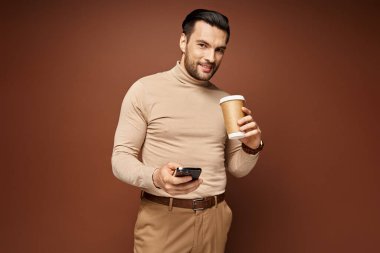 Balıkçı yaka giyen, kahve içen ve telefonuyla bej arka plan, sosyal medya kullanan mutlu adam.