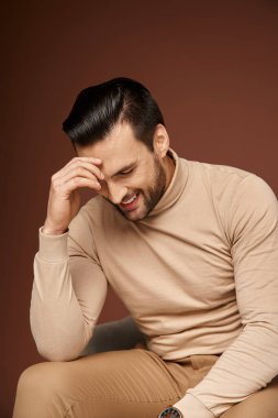 Balıkçı yakalı ve yakışıklı bir adam rahat koltukta oturuyor ve bej arka planda gülümsüyor.