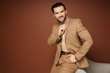 Şık takım elbiseli yakışıklı adam bej arka planda koltuğun yanında el ele poz veriyor.
