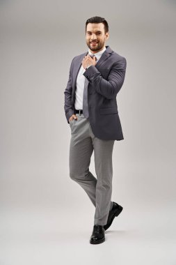 Şık takım elbiseli mutlu adam kravatını düzeltiyor ve gri arka planda el ele poz veriyor.