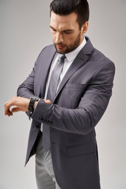 Parlak takım elbiseli yakışıklı iş adamı gri arka planda kol saatini kontrol ediyor.
