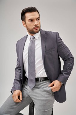 Yakışıklı ve kendine güvenen resmi iş adamı gri arka plandaki yüksek tabureye yaslanıyor.