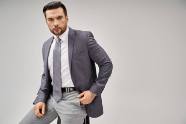 Yakışıklı ve kendine güvenen resmi iş adamı gri arka planda yüksek taburede oturuyor.