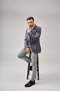 handsome and confident businessman in formal wear leaning on high stool on grey background, elegance clipart