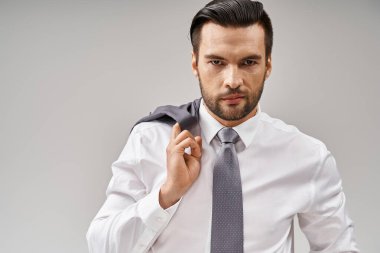 portrait of businessman in his 30s with bristle holding jacket over shoulder while standing on grey clipart