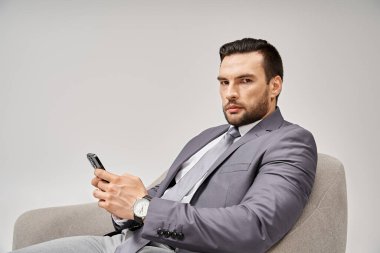 Tüylü iş adamı koltukta oturuyor ve gri arka planda akıllı telefon kullanıyor.