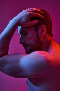 Tüylü seksi ve kaslı bir adam. Mor arka planda eli havada poz veriyor. Üstsüz.