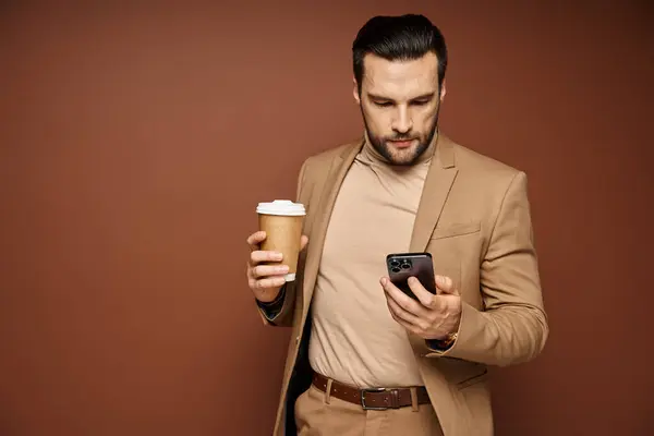 ベージュの背景に彼のスマートフォンを使用して行くためにコーヒーを保持するスタイリッシュな服装のハンサム男 — ストック写真