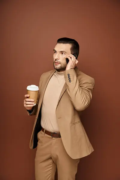 ベージュバックドロップでスマートフォンに電話して行くためにコーヒーを保持するスタイリッシュな服装のハンサム男 — ストック写真