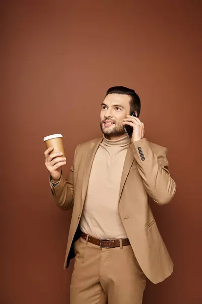 ベージュの背景にスマートフォンで通話するためにコーヒーを保持するスタイリッシュな服装のコンテンツ男 — ストック写真