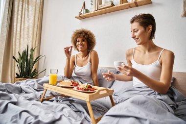 Mutlu, çok kültürlü lezbiyen çift sabah vakti yatakta kahvaltı, içki ve yemek yiyor.