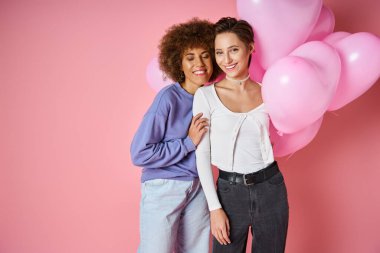 Valentines day concept, joyful multicultural lesbian couple smiling near heart shaped balloons clipart