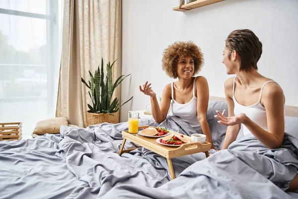 stock image happy multicultural lesbian couple in pajamas gesturing and chatting near breakfast in bed