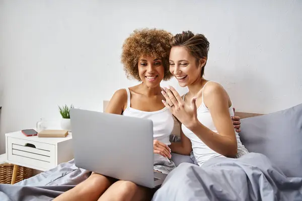 happy engaged woman showing ring on finger during video call near curly african american fiancee