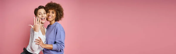 stock image happy lesbian woman showing engagement ring on her finger near female partner on pink, banner