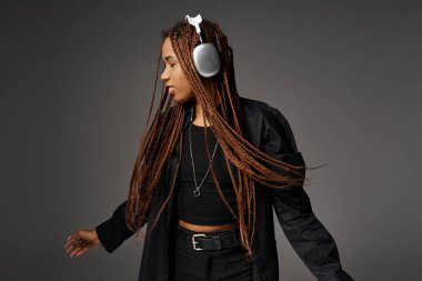 Rahat ve 20 'li yaşlarda, kulaklıklı, gri arka planda müzik eşliğinde dans eden Afro-Amerikan bir kadın.