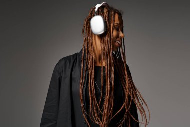 joyful and young African American woman in 20s with headphones listening to music on grey backdrop clipart